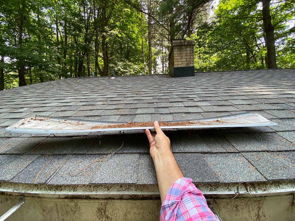 holding a warped 5 foot section of leaffilter gutter guard. the gutter guard appears to be smiling because it is warped upwards at both ends and dips down towards the middle, just like a smile. it still has pine needles sitting on top of the micro mesh screen